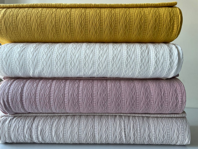 May include: Four folded blankets in a stack, each in a different colour. The top blanket is mustard yellow, followed by white, pale purple, and light grey. Each blanket features a textured cable-knit design.