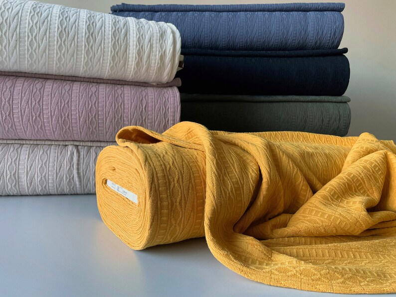 May include: A selection of folded fabric in shades of cream, lilac, and navy. A roll of yellow fabric is in the foreground. The fabric displays a textured, woven design.