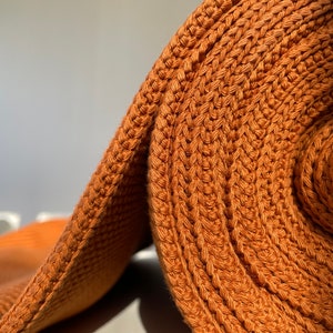 May include: A close-up of a roll of rust-colored crocheted fabric. The fabric is made of a thick, textured yarn and has a slightly bumpy surface.