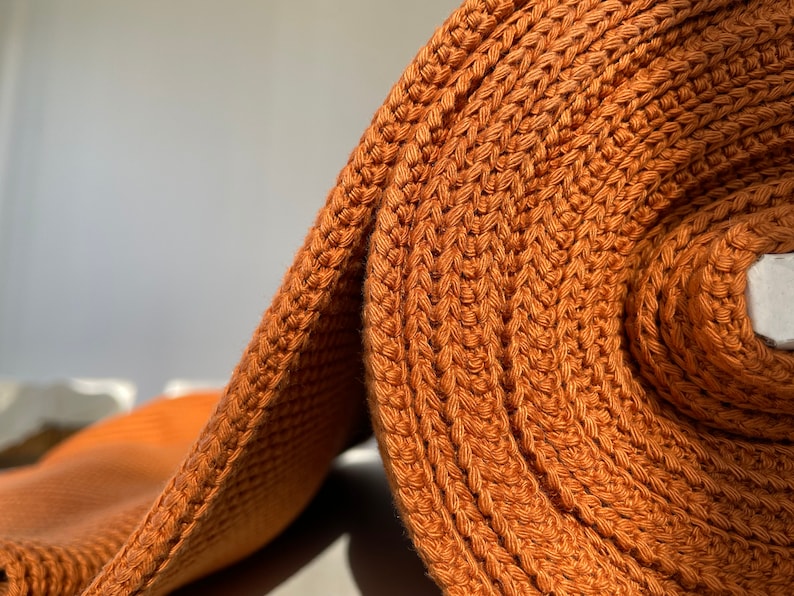 May include: A close-up of a roll of rust-colored crocheted fabric. The fabric is made of a thick, textured yarn and has a slightly bumpy surface.