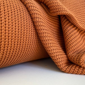 May include: Close-up of a brown knitted fabric with a textured pattern. The fabric is folded and rolled, showing the texture and weave.