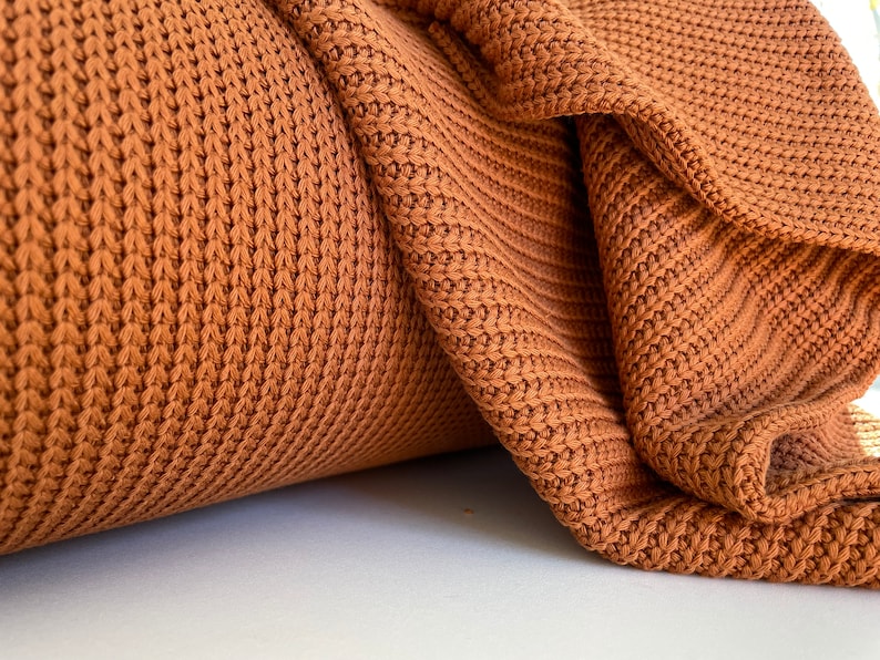 May include: Close-up of a brown knitted fabric with a textured pattern. The fabric is folded and rolled, showing the texture and weave.