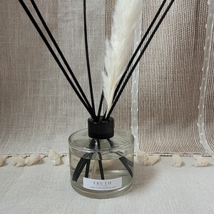 May include: A clear glass diffuser bottle with black reeds and a white fluffy plume. The bottle has a black cap and a label that reads "TRUTH bodyworks". The background is a neutral-toned fabric.
