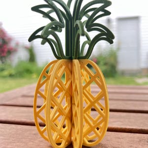 May include: A yellow and green 3D printed pineapple ornament. The pineapple is made of a lattice design and has a green leafy top.