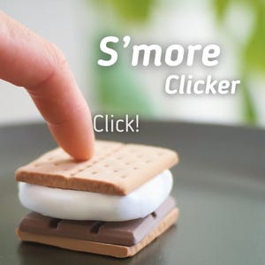 Op de afbeelding: Een s'more-vormige stressbal met een bruine graham cracker, een witte marshmallow en een chocolade reep. De tekst "S'more Clicker" en "Click!" staat op de afbeelding.