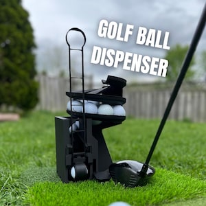 Puede incluir: Un dispensador de pelotas de golf negro con el texto "GOLF BALL DISPENSER" en blanco. El dispensador contiene varias pelotas de golf blancas y está colocado sobre hierba verde. Un palo de golf está en primer plano.