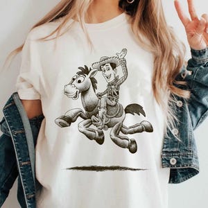May include: Cream-colored t-shirt featuring a black and white illustration of a cowboy riding a horse. The cowboy is wearing a hat and waving. The horse is in mid-gallop. The design is centered on the shirt.