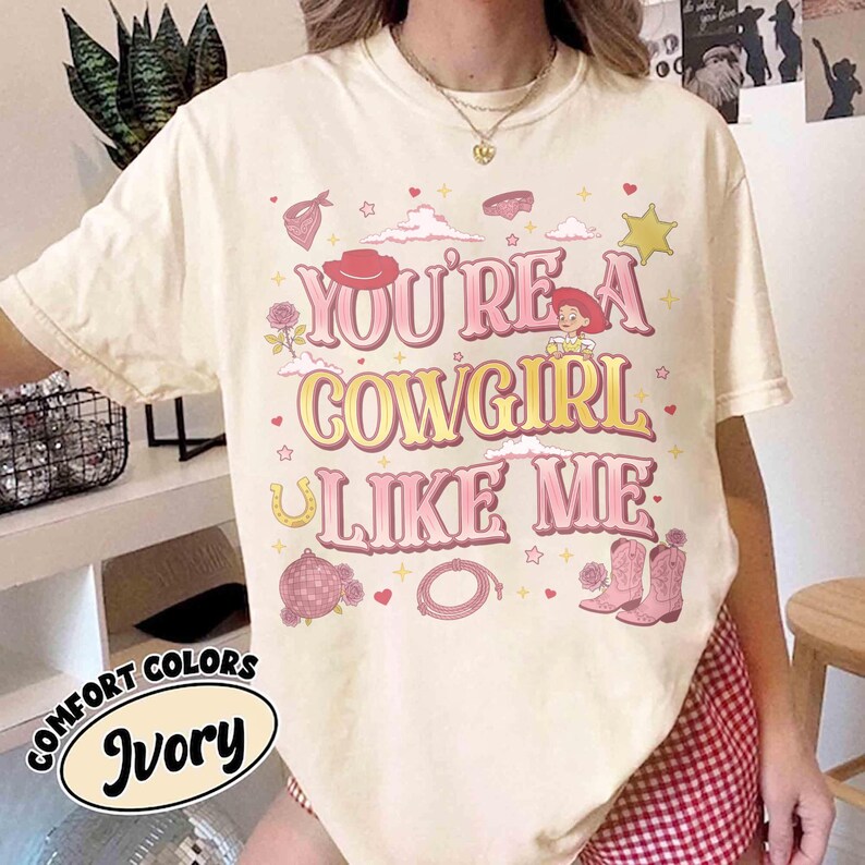 May include: An ivory short-sleeve t-shirt featuring the text "YOU'RE A COWGIRL LIKE ME" in pink and yellow. The graphic includes illustrations of a cowgirl character, pink cowboy boots, a lasso, a disco ball, a horseshoe, a bandana, and stars.