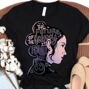 Puede incluir: Camiseta negra con el perfil de una mujer y el texto "THE FUTURE OF THE GALAXY IS FEMALE" en degradado de morado y azul. El diseño incluye estrellas y un símbolo.