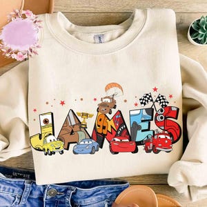 May include: Cream-coloured sweatshirt with the name "JAMES" in a colourful cartoon car design. The design includes various car characters, chequered flags, and a parachute. A fun and playful design for children.