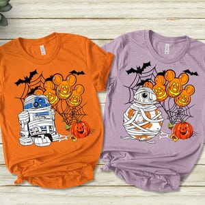 Puede incluir: Dos camisetas con temática de Halloween. Una es naranja con un gráfico de R2-D2 como momia, calabazas, murciélagos y telarañas. La otra es morada con un BB-8 momificado, calabazas, murciélagos y telarañas. Ambas camisetas tienen cuello redondo.