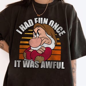 May include: Dark brown t-shirt featuring a graphic of Grumpy from Snow White with the text "I had fun once, it was awful." The design includes a sunset background with orange and yellow stripes.