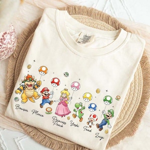 以下が含まれることがあります： クリーム色のTシャツ。ビデオゲームのキャラクターのグラフィックが描かれています。デザインには、クッパ、マリオ、ピーチ姫、ヨッシー、キノピオ、ルイージが含まれており、それぞれがキノコの風船を持っています。キャラクターの名前が下に印刷されています。