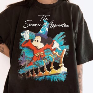 May include: Black t-shirt featuring a graphic of Mickey Mouse as The Sorcerer's Apprentice. Mickey is wearing a blue wizard hat and red robe, with the text "The Sorcerer's Apprentice" above the image.