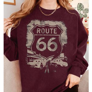 May include: A maroon long-sleeve sweatshirt featuring a distressed graphic of a Route 66 road sign. The design includes two cartoon-style cars positioned below the sign, with the text Route 66 clearly visible in the center of the shield-shaped emblem.