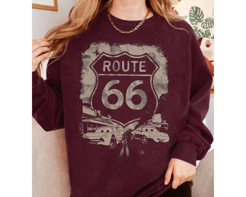 May include: A maroon long-sleeve sweatshirt featuring a distressed graphic of a Route 66 road sign. The design includes two cartoon-style cars positioned below the sign, with the text Route 66 clearly visible in the center of the shield-shaped emblem.