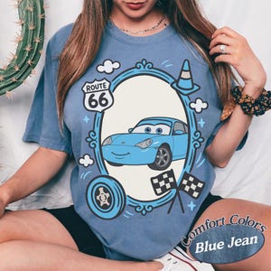 May include: Blue t-shirt with a cartoon car design inside an oval frame, featuring Route 66 signage, clouds, and racing flags. The shirt is labeled "Comfort Colors Blue Jean".