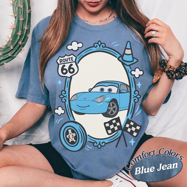May include: Blue t-shirt with a cartoon car design inside an oval frame, featuring Route 66 signage, clouds, and racing flags. The shirt is labeled "Comfort Colors Blue Jean".