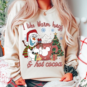 Pode incluir: Camisola bege com um gráfico festivo. Apresenta Olaf, o boneco de neve, uma árvore de Natal e uma caneca de chocolate quente. O texto diz "I like Warm hugs & hot cocoa". O design inclui flocos de neve e caixas de presente.