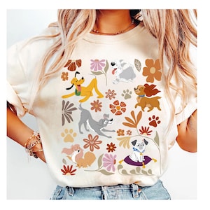 May include: Cream-colored t-shirt featuring a colorful pattern of cartoon dogs, flowers, and paw prints. The dogs are in various poses, with a mix of colors and styles. The floral elements include brown, pink, and white flowers. The shirt is a casual, everyday wear item.