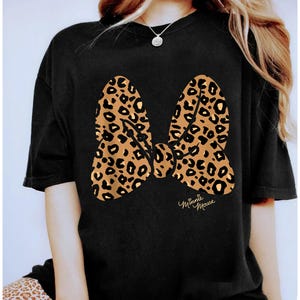 Op de afbeelding: Zwart T-shirt met een groot luipaardprint strik ontwerp. De strik is in bruin- en beigetinten met zwarte stippen. De woorden "Minnie Mouse" staan eronder in cursief.