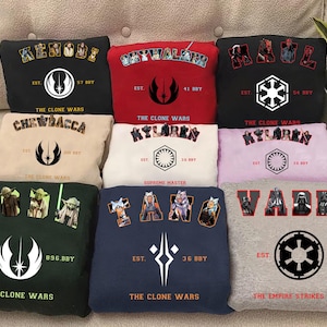 May include: A selection of Star Wars themed sweatshirts in colours such as black, red, and grey. Each sweatshirt displays character names, logos, and text from the Star Wars franchise. The sweatshirts are arranged on a beige surface.