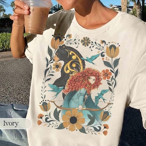Puede incluir: Camiseta de color marfil con una ilustración colorida. El diseño incluye un oso negro, una mujer con cabello rizado rojo, pájaros azules, flores y un arco y flecha. La palabra "Ivory" está impresa en la parte inferior izquierda.