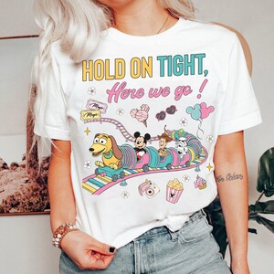 Op de afbeelding: Wit T-shirt met een kleurrijke afbeelding van een cartoon trein met personages en de tekst "HOLD ON TIGHT, Here we go!". Het ontwerp bevat Mickey Mouse, Woody en een Stormtrooper.