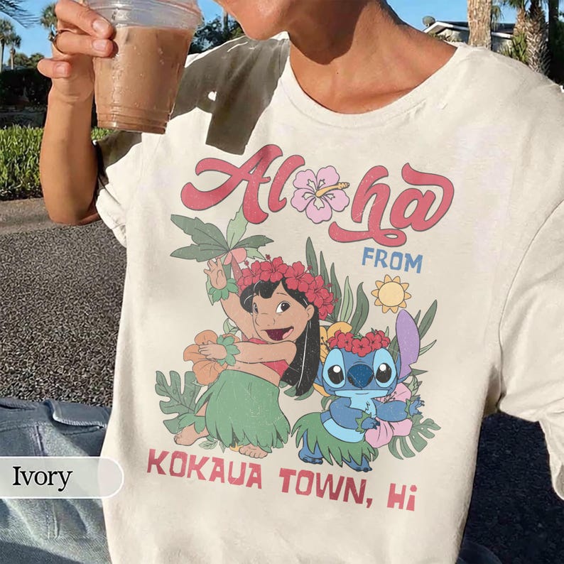 May include: Ivory-colored t-shirt featuring the text "Aloha From Kokaua Town, Hi" in red and pink. The graphic includes cartoon characters, flowers, and leaves. The shirt is a casual wear item.