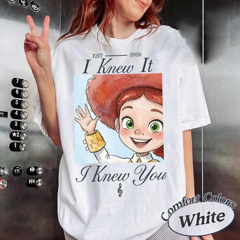 Peut inclure: Un t-shirt blanc avec une illustration d'un personnage de dessin anim&eacute; aux cheveux roux portant un grand chapeau rouge et faisant un signe de la main. Le texte indique EST 2026, I Knew It, et I Knew You, avec une note de musique sous le texte.