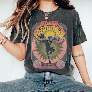 Könnte beinhalten: Dunkelgraues T-Shirt mit einem Retro-Grafikdesign von Black Panther. Das Design zeigt den Text "Black Panther" in einer geschwungenen Schrift, eine Panther-Illustration und die Worte "Wakanda Forever". Das T-Shirt hat einen Vintage-Look.