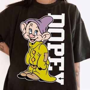 May include: Black t-shirt featuring a cartoon image of Dopey from Snow White, wearing a yellow outfit and purple hat. The word "DOPEY" is printed vertically in white block letters on the right side of the shirt.