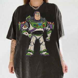 May include: A charcoal black short-sleeve t-shirt featuring a large, centered graphic print of the character Buzz Lightyear. The character is depicted in a white, green, and purple space suit with wings extended, standing in a confident pose.