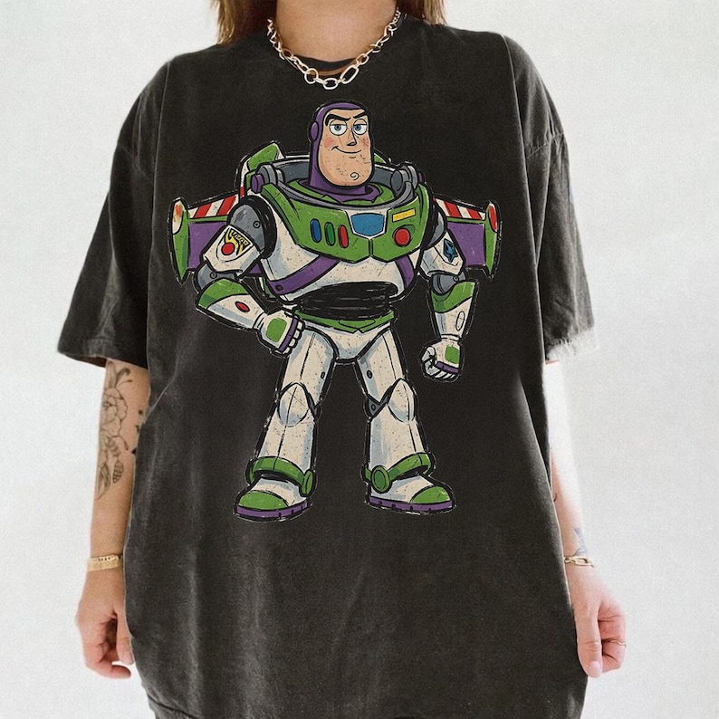 May include: A charcoal black short-sleeve t-shirt featuring a large, centered graphic print of the character Buzz Lightyear. The character is depicted in a white, green, and purple space suit with wings extended, standing in a confident pose.
