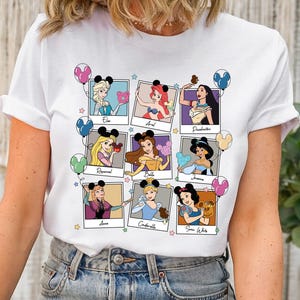 Può includere: T-shirt bianco con un collage di nove principesse Disney in cornici in stile polaroid. Ogni principessa è raffigurata con una fascia per capelli con orecchie di Topolino e un palloncino. Le principesse includono Elsa, Ariel, Pocahontas, Rapunzel, Belle, Jasmine, Anna, Cenerentola e Biancaneve.