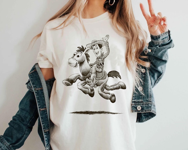 May include: Cream-colored t-shirt featuring a black and white illustration of a cowboy riding a horse. The cowboy is wearing a hat and waving. The horse is in mid-gallop. The design is centered on the shirt.