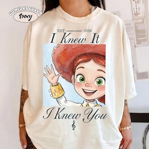 Peut inclure: Un t-shirt ivoire avec une illustration style aquarelle d'un personnage aux cheveux roux portant un grand chapeau rouge, des yeux verts et une manchette jaune. Le texte indique "EST 2026 I Knew It I Knew You" avec une note de musique.