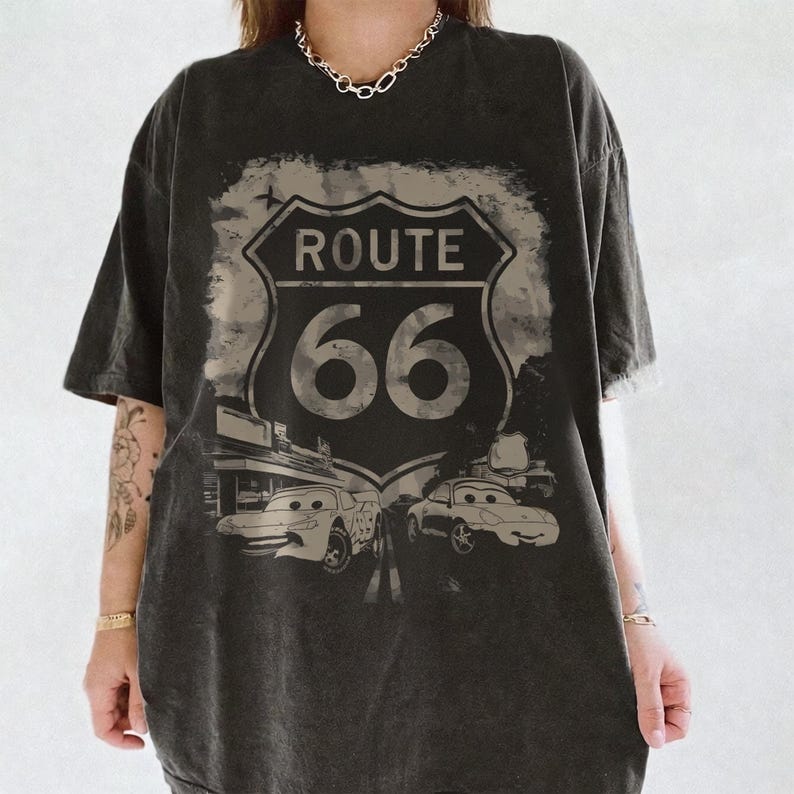 May include: A charcoal grey oversized t-shirt featuring a distressed graphic of the Route 66 highway sign. Below the sign, two cartoon-style race cars are depicted on a road. The shirt has short sleeves and a crew neckline, with a faded, vintage look.