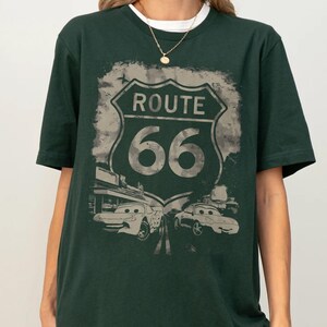 May include: A dark green short-sleeve t-shirt featuring a large graphic print of a Route 66 road sign. Below the sign, the design includes two cartoon-style cars with eyes on their windshields, set against a distressed, muted background.