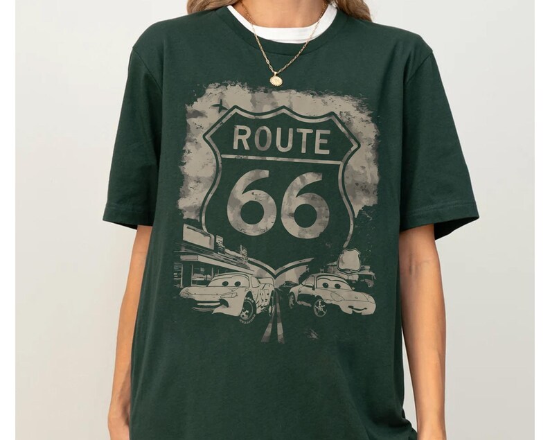May include: A dark green short-sleeve t-shirt featuring a large graphic print of a Route 66 road sign. Below the sign, the design includes two cartoon-style cars with eyes on their windshields, set against a distressed, muted background.