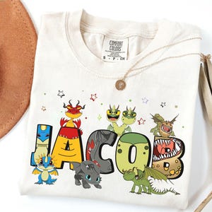 Puede incluir: Camiseta color crema con el nombre "JACOB" en letras coloridas de estilo caricatura, cada una adornada con una ilustración de dragón diferente. La camiseta tiene cuello redondo y mangas cortas. Pequeñas estrellas y puntos están dispersos alrededor del nombre.