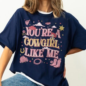May include: A navy blue t-shirt featuring a graphic print with the text "YOU'RE A COWGIRL LIKE ME" in pink and yellow. The design includes illustrations of a cowgirl character, pink cowboy boots, a lasso, a disco ball, a horseshoe, and stars.