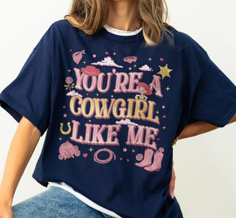 May include: A navy blue t-shirt featuring a graphic print with the text "YOU'RE A COWGIRL LIKE ME" in pink and yellow. The design includes illustrations of a cowgirl character, pink cowboy boots, a lasso, a disco ball, a horseshoe, and stars.
