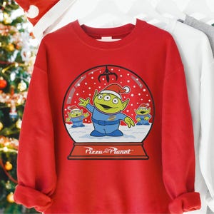 Puede incluir: Una sudadera roja con un diseño de bola de nieve con tres extraterrestres verdes con gorros de Papá Noel. La bola de nieve tiene el texto "Pizza Planet" en la parte inferior. Los extraterrestres son de la franquicia Toy Story.