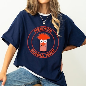 May include: Navy blue t-shirt with a white collar featuring a graphic of a Muppet character inside a red circle. The text "MEEPERS GONNA MEEP" is printed around the character.