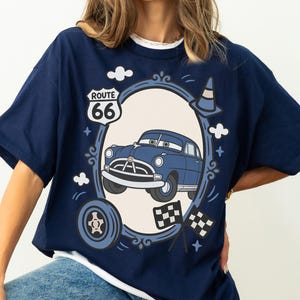May include: Navy blue t-shirt with a cartoon car design inside an oval frame. The design includes a Route 66 sign, clouds, a traffic cone, a tyre, and checkered flags. The car is blue with white detailing.
