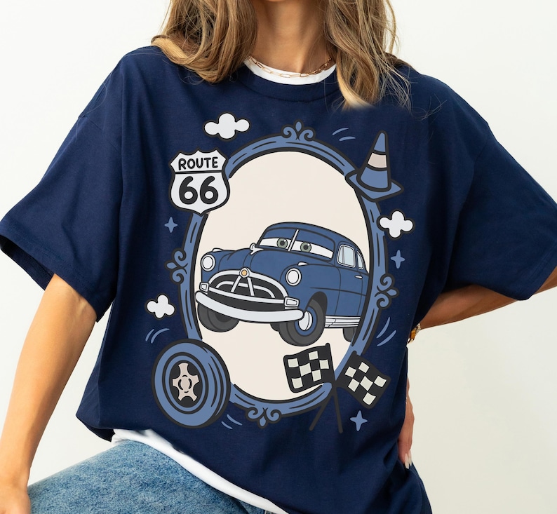 May include: Navy blue t-shirt with a cartoon car design inside an oval frame. The design includes a Route 66 sign, clouds, a traffic cone, a tyre, and checkered flags. The car is blue with white detailing.