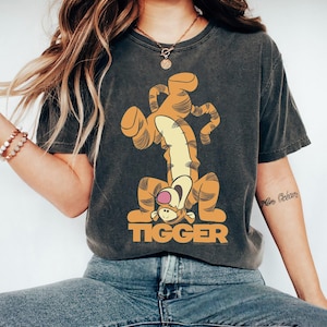 Puede incluir: Camiseta gris oscuro con una imagen de dibujos animados de Tigger de Winnie the Pooh. El Tigger naranja y amarillo está boca abajo con la palabra "TIGGER" en letras naranjas debajo. La camiseta tiene un aspecto vintage y desgastado.