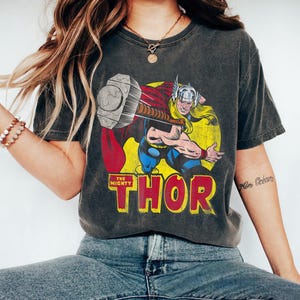 May include: Dark gray t-shirt featuring a vintage comic book illustration of Thor, wielding his hammer. The graphic includes the text "THE MIGHTY THOR" in yellow and red. The shirt has a classic, worn look.