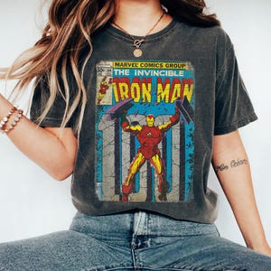 May include: Dark gray t-shirt featuring a vintage comic book cover of Iron Man. The comic book cover has a red, yellow, and blue color scheme with the text "Iron Man" in large yellow letters. The shirt is worn by a person.
